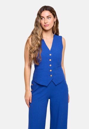 Mujer con cabello largo y ondulado que lleva un chaleco azul sin mangas con botones y pantalones azules a juego con detalles de botones en el lateral.