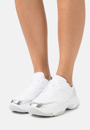 Baskets blanches épaisses avec des accents texturés argentés portées aux pieds sur un fond clair uni.