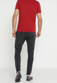 Camisa roja de manga corta y pantalones de chándal oscuros ajustados en gris. La tela parece suave; los pantalones tienen puños en los dobladillos. El calzado presenta una mezcla de colores.