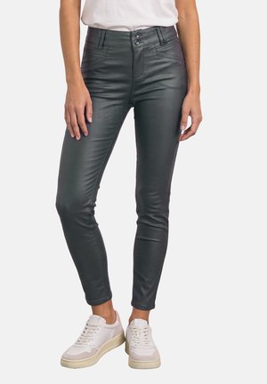 Femme portant un pantalon en cuir noir slim et des baskets blanches, debout devant un fond blanc uni.