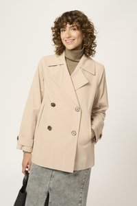 Chaqueta beige de corte cruzado con grandes botones, solapas anchas y un ajuste relajado. Lleva puesta una camiseta de cuello alto gris y se combina con una falda gris.