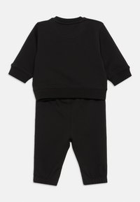 Μαύρη σύνολο φούτερ και παντελονιού φτιαγμένο από μαλακό ύφασμα. Το πουκάμισο έχει ραμμένες μανσέτες; το παντελόνι διαθέτει ελαστική μέση και φαρδιά πόδια.
