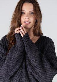 Donkergrijs geribbeld gebreid sweater met een V-hals, oversized mouwen en textuurstof, met vingers die zachtjes de mouwen vasthouden.