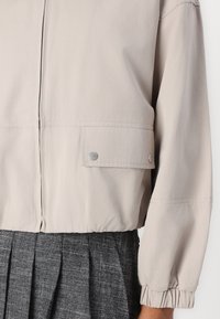 Chaqueta beige claro con cierre de cremallera, puños elásticos y un bolsillo con botón a presión. El material parece suave y con un ligero brillo.