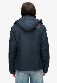Granatowa ocieplana kurtka z kapturem, zapinana na zamek i napy, z akcentami logo na ramieniu i rękawie. Teksturowana tkanina.