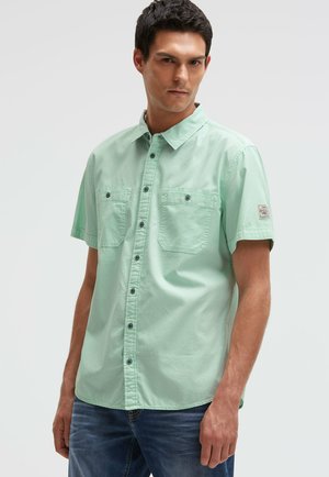 Mann trägt ein hellgrünes kurzärmeliges Hemd mit zwei Brusttaschen und blaue Jeans vor einem einfarbigen Hintergrund.