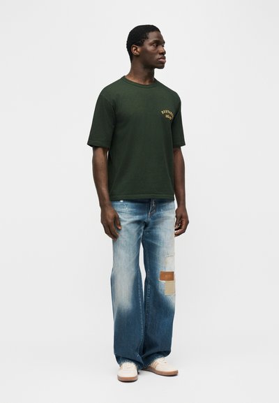 Άνδρας που φοράει σκούρο πράσινο κοντομάνικο μπλουζάκι με κίτρινα γράμματα και ξεβαμμένο τζιν φαρδιάς γραμμής με μπαλώματα, στέκεται μπροστά σε ένα απλό ανοιχτό φόντο.