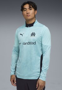 Tee-shirt de sport à manches longues bleu clair avec un col zippé mi-haut, des accents noirs, un logo sur la poitrine et un motif texturé. Fabriqué en tissu de sport.