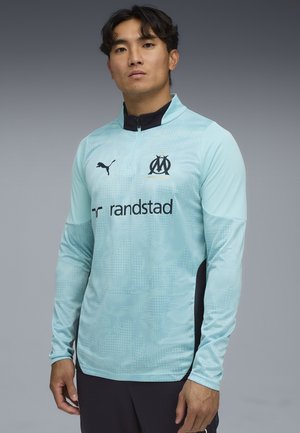 Tee-shirt de sport à manches longues bleu clair avec un col zippé mi-haut, des accents noirs, un logo sur la poitrine et un motif texturé. Fabriqué en tissu de sport.