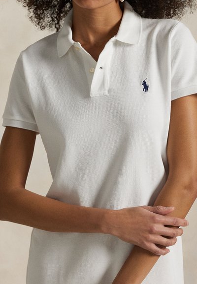 Valkoinen pikeepaita, jossa on teksturoitu kangas, lyhyet hihat ja käännetty kaulus. Vasemmassa rinnassa pieni tummansininen logo.