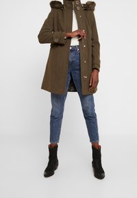 Persona vistiendo un abrigo verde oliva con capucha de piel sintética, jeans azules, botas negras hasta el tobillo y una camisa blanca, de pie contra un fondo liso.