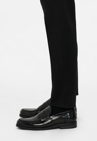 Mustad musta nahast loafers, millel on poleeritud viimistlus, õmblustega detail ja lame tall, kombineeritud mustade pükstega.