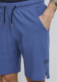 Pantaloni corti blu con texture, dotati di vita elastica, cordoncino e tasche laterali. Un'etichetta è visibile sulla gamba sinistra.