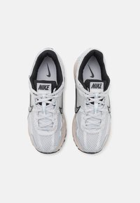 Nike Vomero-sneakers har en grå mesh-överdel med en svart inredning och en beige sula. Skosnörerna är vita med en subtil blå accent.