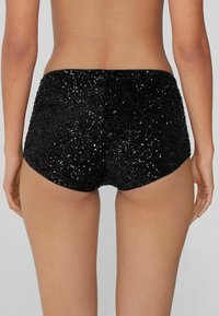 Pantalones cortos negros con una superficie texturizada y de lentejuelas, que presentan un diseño ajustado y bordes suaves, mostrando un patrón detallado y brillante.