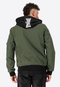 Olivovo zelená bomber bunda s čiernou kapucňou. Má biele potlačené logo na kapucni, elastické manžety a voľný strih.