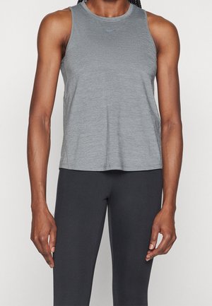 Osoba v šedém sportovním tílku a černých legínách stojí před světlým jednobarevným pozadím.