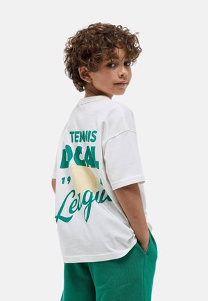Jeune garçon aux cheveux bouclés portant un tee-shirt de tennis blanc oversize avec un texte vert et un short bleu sarcelle, les mains dans les poches, regardant par-dessus l'épaule.