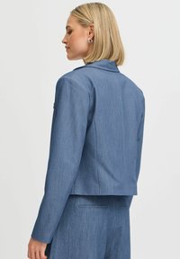 Kék denim hatású dzseki, testhezálló fazonnal, sima textúrával, kivágott gallérral és hosszú ujjakkal, cropped derékkal és minimális varrásokkal.