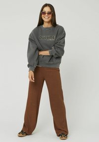 Sweatshirt gris avec impression "OUTSIDE", coupe large, épaules tombantes, associé à un pantalon large marron et des chaussures imprimé léopard. Modèle souriant.