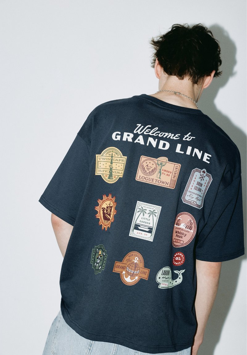 Personne portant un t-shirt noir avec le texte "Welcome to Grand Line" et neuf dessins de badges de voyage colorés dans le dos.