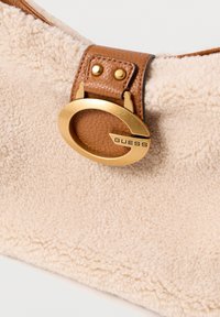 Hnědá kabelka z umělého beránka s hnědým koženým popruhem. Obsahuje zlatý kruhový kovový prvek s nápisem "GUESS" pro detail.