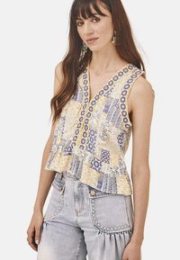 Top sin mangas blanco con un diseño de patchwork en amarillo y azul, con un escote en V y un dobladillo con volantes. Combinado con jeans de mezclilla claros.