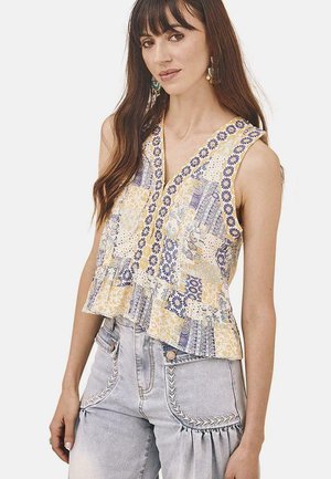 Top sin mangas blanco con un diseño de patchwork en amarillo y azul, con un escote en V y un dobladillo con volantes. Combinado con jeans de mezclilla claros.