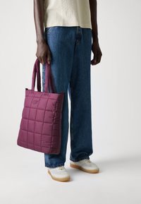 Quiltet bordeaux håndveske med doble håndtak, laget av polstret materiale, vist mot en nøytral bakgrunn, sammen med blå jeans og joggesko.