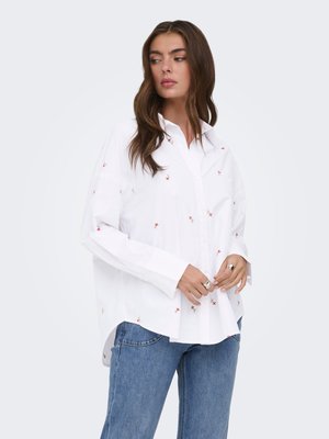 Sieva ar gariem brūniem matiem, ģērbta baltā pogājama kreklā ar mazām sarkanām ziedu rakstām un zilās džinsās, skatās uz sāniem, rokas savilktas kopā.