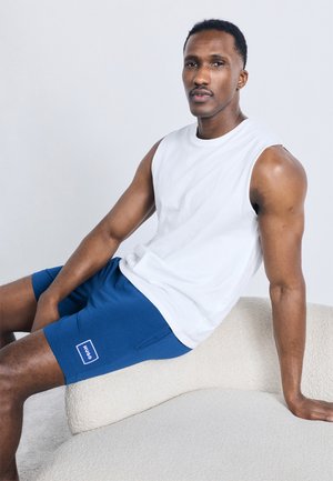 Homme portant un débardeur blanc et un short bleu Hugo assis sur une chaise texturée de couleur crème sur un fond gris.