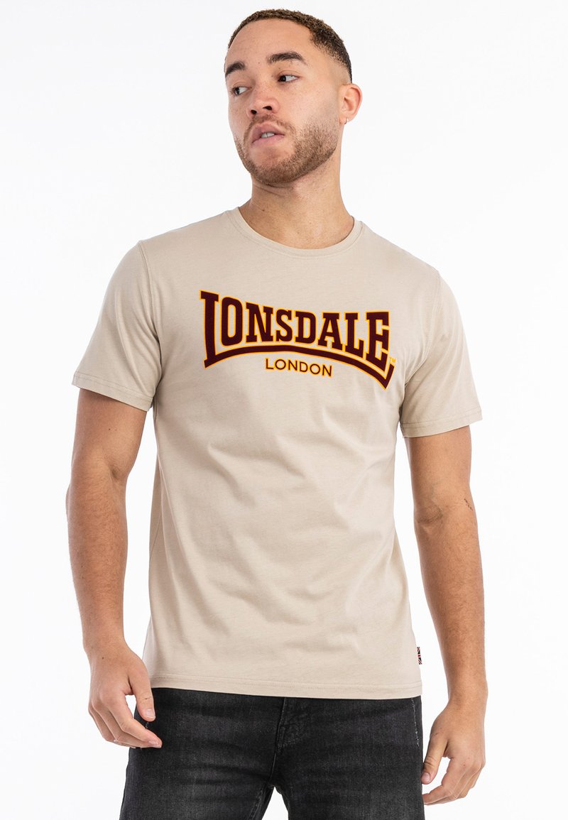 Smėlio spalvos medvilnės marškinėliai su dideliu tamsiai raudonu "Lonsdale London" logotipu per krūtinę. Klasikinis apvalus kaklas ir trumpi rankoviai.