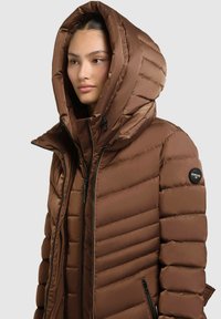 Chaqueta de plumas marrón con una gran capucha. Presenta una textura acolchada horizontal, dos cremalleras negras y un parche circular de la marca en la manga.