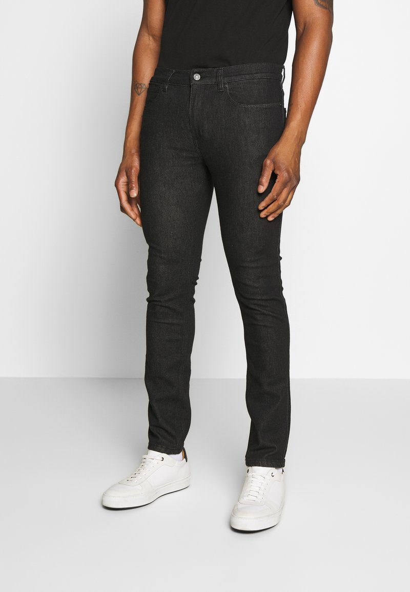 Vaqueros skinny negros de denim, con un clásico diseño de cinco bolsillos y cierre de botón. Combinados con zapatillas blancas.