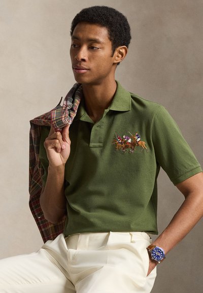 Άνδρας που φοράει ένα ελιά-πράσινο πόλο με κεντημένους παίκτες πόλο, κρεμ παντελόνι, ένα καφέ ρολόι και κρατάει ένα καρό σακάκι στον ώμο του.