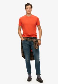 Orange kortærmet t-shirt, blå jeans og en brun rutet skjorte bundet om taljen. Accessories inkluderer et sort bælte og mørke sko.