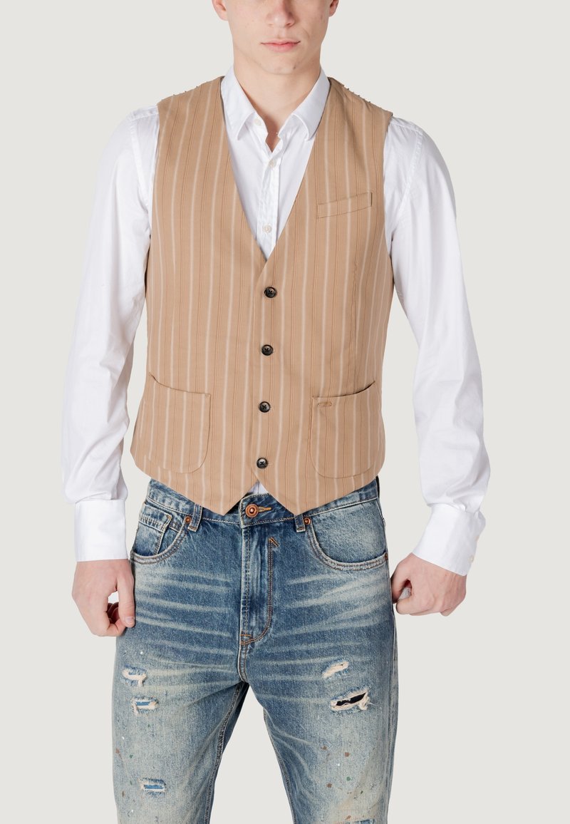 Gilet a righe beige con cinque bottoni e tasche, indossato sopra una camicia bianca, abbinato a jeans blu strappati con macchie di vernice.