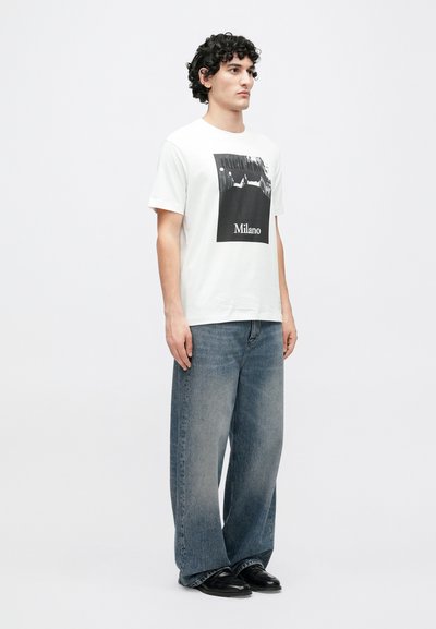 Biele tričko s čiernym grafickým motívom s textom "Milano". Kombinované s voľnými svetlomodrými džínsami a čiernymi topánkami. Hladká textúra látky.