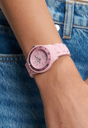 Lyserødt analogt armbåndsur med silikonerem, båret på en persons venstre arm over blå denimjeans.