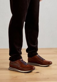 Zapatillas de cuero marrón con suela de goma blanca y gris, cordones azul marino y acentos laterales. Llevadas con pantalones de pana oscuros sobre un suelo de madera.