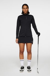 Sort tætsiddende langærmet zip-jakke kombineret med en matchende nederdel, sorte handsker og sort-hvide sportssko, mens der holdes en golfkølle.