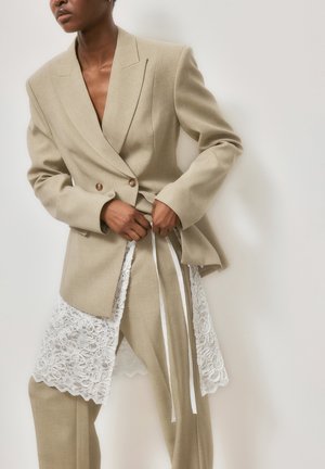 Ženska v bež prilagojenem kostimu z belim čipkastim robom, ki si zavezuje pas proti svetlemu ozadju.