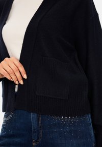 Dunkelblauer Cardigan mit offenem Vorderteil, geripptem Saum und einer aufgesetzten Tasche; über einem weißen Top getragen und mit verzierten Jeans kombiniert.