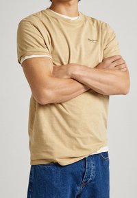Beige bomulls-t-shirt med rundad halsringning, kontrasterande vit kant och en liten logotyp på vänster bröst; kombinerad med blå denimjeans.