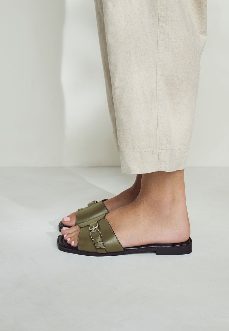 Sandales mules vert olive avec une large bride, un détail de boucle en métal et une semelle noire plate. Le pied est partiellement visible, positionné de côté.