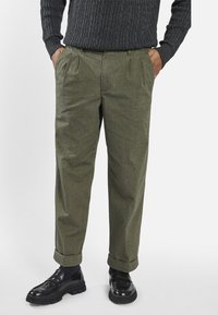 Persoon in olijfgroene geplisseerde broek met manchetten, zwarte chunky loafers, donkere sokken en een donkergroen kabelgebreid sweater, met de handen in de zakken.
