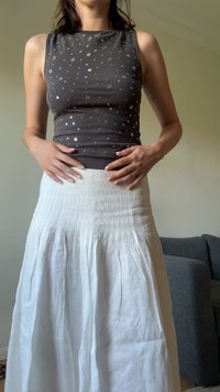 Grijze mouwloze top met een metallic stippenpatroon, gecombineerd met een witte geplooide rok. De stof lijkt licht en gestructureerd.