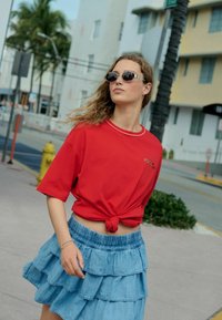 Rød kortærmet skjorte knyttet i taljen, parret med en lagdelt denimnederdel med volangdesign. Afsluttet med runde solbriller.