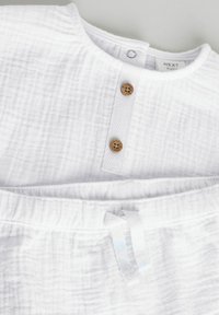 Conjunto de bebé de algodón blanco que incluye una parte superior abotonada con dos botones de madera y pantalones cortos con cintura elástica y cordón. Tejido suave y texturizado.