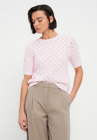 Pull en maille rose clair à manches courtes, avec un motif ajouré décoratif. Associé à un pantalon plissé beige pour un look texturé.
